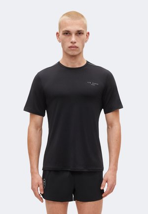 Ung mand med kort blond hår iført en sort Ted Baker Sport t-shirt og sorte shorts mod en ensfarvet lys baggrund.