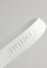 Correa de goma blanca con superficie texturizada y el logotipo "SUPERDRY" en relieve. Forma curvada con bordes suaves. Diseño minimalista.