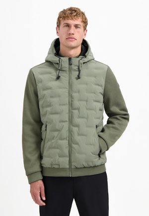 Man in een olijfgroene gewatteerde jas met capuchon en gebreide mouwen, met zwarte broek, staande met één hand in zijn zak tegen een witte achtergrond.