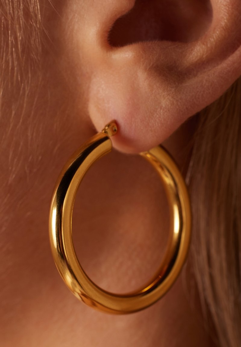 Boucle d'oreille en or avec une finition lisse et polie, présentant une forme arrondie et un design de fermoir sécurisé, portée sur une oreille.