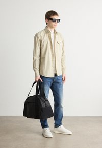 Camicia beige con bottoni, t-shirt bianca sotto, jeans blu, borsa nera con texture di tessuto doppie e sneakers bianche. Occhiali da sole neri indossati.
