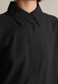 Svart skjorte med spiss krage, seks knapper foran og glatt tekstur. Har svarte knapper og minimal sømdetalj.