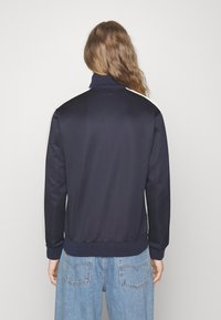 Veste bleu marine en tissu lisse, avec un col montant et des bandes latérales blanches. Poignets et ourlet côtelés. La vue de dos présente un design simple.