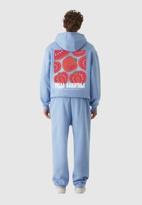 Mėlyna džemperis su vaisių grafika ir tekstu ant nugaros, derinamas su atitinkančiomis mėlynomis kelnių kelnėmis ir baltais sportbačiais. Minkšta, laisvalaikio medžiaga.