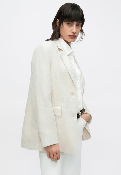 Beiger Leinenblazer mit lockerem Schnitt, einreihiger Knopfleiste, notched Revers und Seitentaschen. Kombiniert mit einem weißen Hemd mit Knopfleiste.