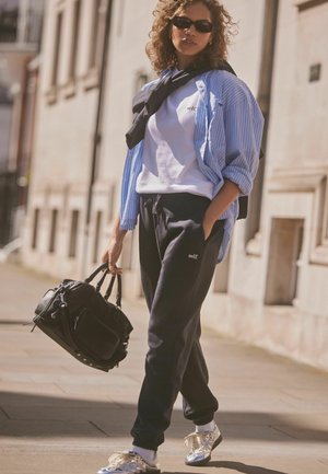 Marineblaue Sweatpants, weißes T-Shirt, übergroßes gestreiftes Hemd, schwarze Handtasche und metallische Sneakers, accessoriert mit Sonnenbrille.