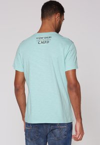 Světle modré bavlněné tričko s kulatým výstřihem. Na horní zadní části má tištěný design. Krátké rukávy a volný střih.