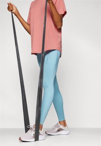 T-shirt sportivo rosa chiaro, leggings blu con cuciture laterali e scarpe da allenamento grigie con suole bianche e dettagli neri; in mano bande di resistenza nere.