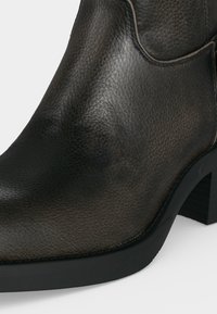 Gros plan d'une botte cheville en cuir noir avec une surface texturée et un talon épais sur fond blanc.