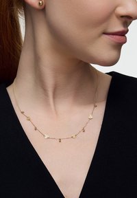 Złoty naszyjnik z małymi zawieszkami w kształcie serca i okrągłymi koralikami na delikatnym łańcuszku. Noszony na czarnej bluzce, ukazujący szczegóły wzoru.