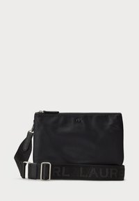 Sac en nylon noir à bandoulière avec fermeture éclair, doté d'une sangle texturée portant le nom de la marque. Forme rectangulaire compacte, surface lisse.