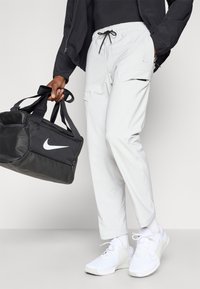 Ανοιχτό γκρι cargo παντελόνι με ελαστική μέση, μαύρο κορδόνι και τσέπες. Μαύρη τσάντα duffel Nike με λογότυπο, σε συνδυασμό με λευκά αθλητικά παπούτσια.