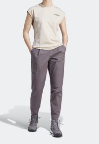 Person klädd i en beige ärmlös Terrex-t-shirt, grå tajta byxor och grå snörskor, stående med händerna i fickorna.