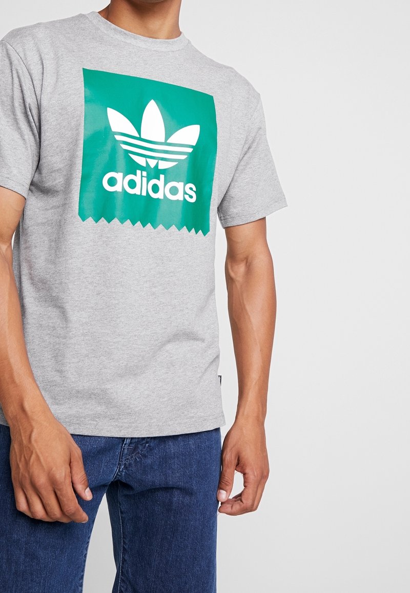 Grå bomullströja med en grön rektangulär grafik som har en vit Adidas-logotyp och tre randiga mönster, korta ärmar och en avslappnad passform.