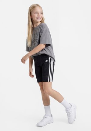 Smilende jente med langt blond hår iført grå oversized t-skjorte, svarte Adidas-shorts med hvite striper, hvite sokker og hvite sneakers, stående.