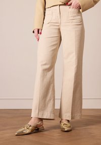 Beige wijdvallende broek in katoenmix, met twee achterzakken en een subtiele textuur, gecombineerd met metallic gouden slingback schoenen.