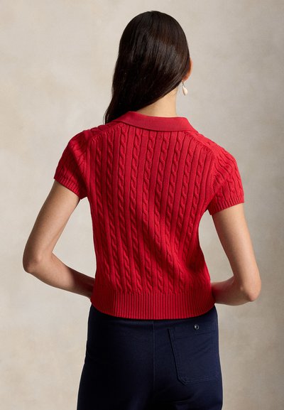 Žena s dlhými tmavými vlasmi, oblečená v červenom krátkym rukávom svetríku s vrkočovým vzorom a tmavých nohaviciach, stojaca chrbtom k hladkému pozadiu.