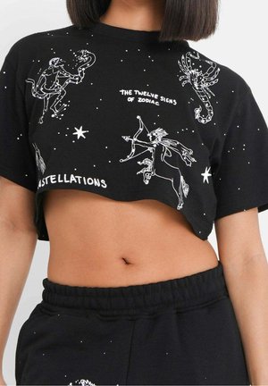T-shirt court noir avec des illustrations blanches de constellations du zodiaque et texte, porté avec un pantalon noir à cordon assorti.