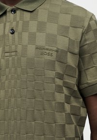 Πόλο πουκάμισο σε ελιά πράσινη απόχρωση με υφασμένο μοτίβο σκακιέρας και ανάγλυφο λογότυπο "PORSCHE BOSS" στο αριστερό στήθος.