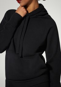 Schwarzer Hoodie aus weichem Baumwollstoff, mit einer Fronttasche, Kapuze mit Kordelzug und gerippten Bündchen für eine bequeme Passform.