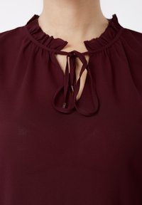 Βυσσινί μπλούζα με γιακά βολάν, άνοιγμα σε σχήμα κλειδαρότρυπας και κορδόνια στο τελείωμα. Ελαφριά υφή με διακριτική γυαλάδα.