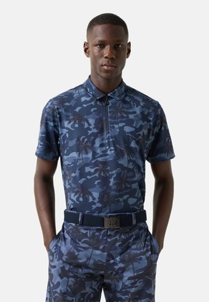 Man wearing matching blue camo-patterned short-sleeve polo shirt and pants with palm tree details and a black belt with a square buckle.