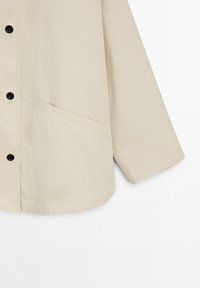 Beige lange mouwen jas met zwarte knopen en een schuine zak aan de linkerkant, getoond tegen een witte achtergrond.