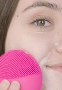 Rosa silikonansiktsrengöring med texturerad yta, hålls mot kinden medan ansiktet tvättas; synligt skum och detaljer av ansiktshuden.
