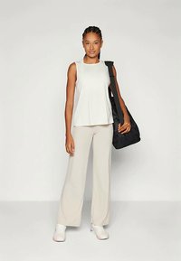 Naine seisab, seljas valge ilma varrukateta top, beežid laiad püksid, valged tossud ja kannab musta õlakotti, halli tausta ees.