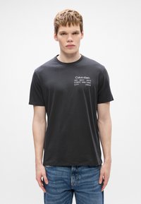 Μαύρη βαμβακερή μπλούζα t-shirt με στρογγυλή λαιμόκοψη, με λευκές λεπτομέρειες κειμένου στο στήθος. Φορεμένη με μπλε τζιν παντελόνι.