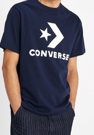 Mężczyzna w granatowym t-shircie z dużym białym logo i napisem Converse, ręce w kieszeniach ciemnych spodni w prążki.