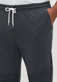 Dark gray sweatpants with white drawstring, textured ribbed detailing on knees, and side pockets with a hand inside one pocket.