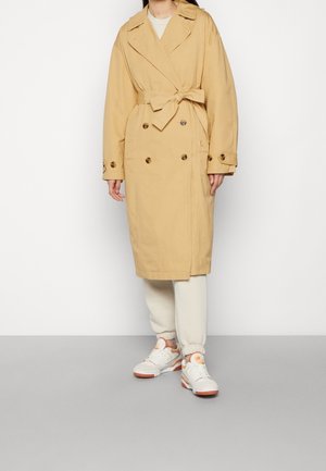 Frau trägt einen beigefarbenen, doppelt geknöpften Trenchcoat mit Taillengürtel, kombiniert mit cremefarbenen Jogginghosen und weißen Sneakers mit orangenen Akzenten.