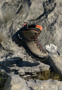 Brązowy but trekkingowy z pomarańczowymi akcentami, teksturowana cholewka i wytrzymała podeszwa, unosi się w powietrzu nad skalistym terenem. Posiada sznurowadła i logo.