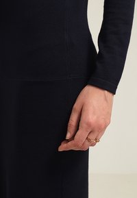 Robe ajustée bleu marine à manches longues, texture côtelée et détails de coutures. Main montrant une bague en or avec un design complexe.