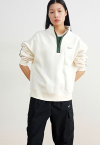 Balts vilnas krekls ar zaļām rāvējslēdzēja apkaklēm un mazu rozā logotipu. Audums šķiet mīksts; tam ir brīva griezuma forma un piedurknes ar aprocēm.