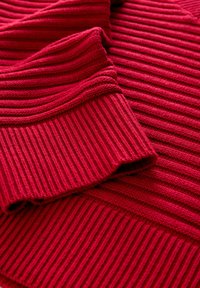 Zbliżenie na czerwony sweter z dzianiny prążkowanej, rękaw złożony na ciele, ukazujący szczegółowe teksturowane szwy i miękkość materiału.