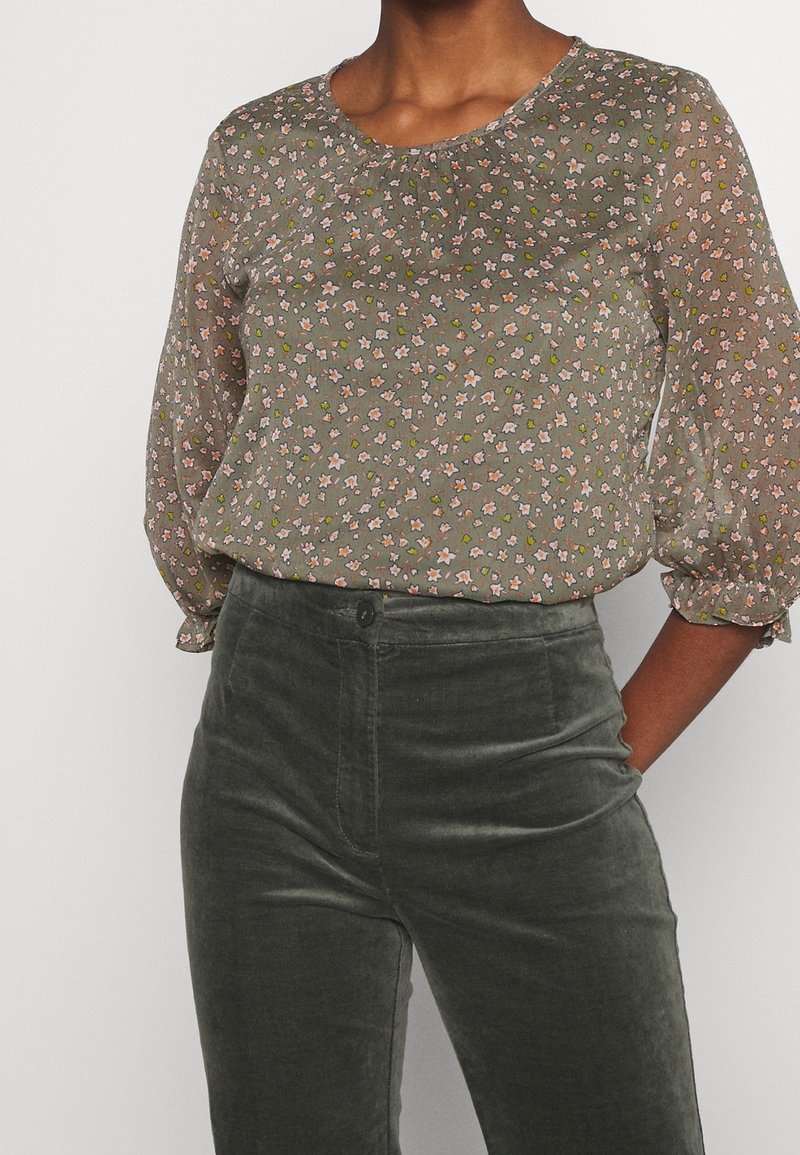 Przezroczysta, oliwkowozielona bluzka z drobnym kwiatowym wzorem, okrągłym dekoltem i marszczeniem na ramionach; zestawiona z ciemnozielonymi, dopasowanymi spodniami.
