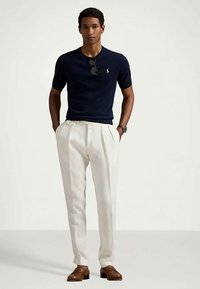 Άνδρας με ναυτικό μπλε κοντομάνικο πουκάμισο, λευκά πλισιέ παντελόνια, καφέ δερμάτινες εσπαντρίγιες, γυαλιά ηλίου στη ζώνη του πουκαμίσου, στέκεται με τα χέρια στις τσέπες.