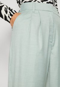 Lichtblauwe geplooide broek met een gladde textuur, met zijzakken en een tailleband. Een zwart-wit gestreepte top is deels zichtbaar.