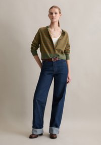 Vrouw staat en draagt een groen-bruine gestreepte cardigan, een wit tanktop, wijd uitlopende opgerolde jeans, een bruine riem en bruine laarzen.