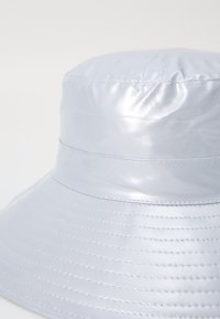 Ljusblå, glansig bucket hat gjord av ett slätt material; har sydda detaljer runt brätten och en subtil söm på toppen.