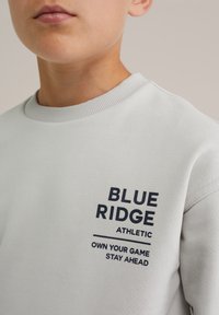 Svetlo šedý sveter vyrobený z bavlneného zmesi, s námorníckym modrým textom: "BLUE RIDGE ATHLETIC" a sloganom pod ním. Žebrovaný golier a manžety.