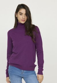 Un pull à col roulé violet en tissu doux, doté de manches longues et d'un ourlet côtelé. Porté avec un jean bleu. Design minimaliste.