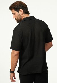 Schwarzes Kurzarmhemd aus strukturiertem Stoff, mit Kragen und lockerer Passform. Sichtbare Nähte entlang der Schultern.