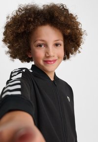 Giacca nera con zip e dettagli a strisce bianche sulle maniche. Tessuto morbido, design aderente e una patch con logo sulla parte frontale.