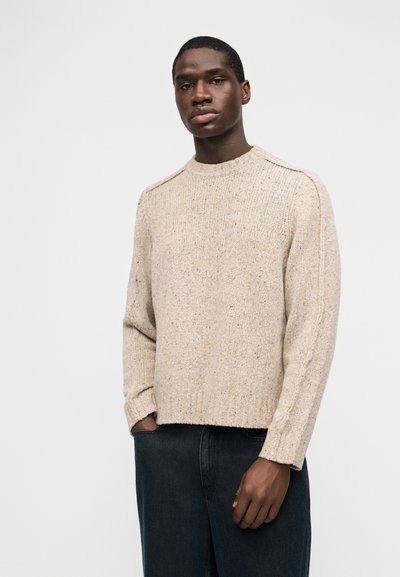 Άνδρας που φοράει μπεζ πλεκτό πουλόβερ και σκούρο τζιν, στέκεται με το ένα χέρι στην τσέπη μπροστά σε απλό λευκό φόντο.