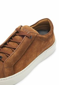 Ruskeahihaiset tennarit, joissa on sävysovitettu nauhoitus, valkoinen kumipohja ja kohokuvioitu logotieto sivussa. Suora sisävuori mukavuutta varten.