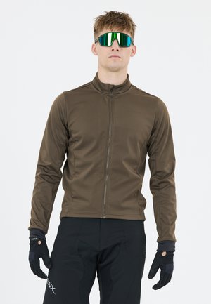 Mann mit grünen verspiegelten Sonnenbrillen, brauner Zip-up-Jacke, schwarzen Handschuhen und schwarzen Hosen vor einem einfarbigen hellen Hintergrund.