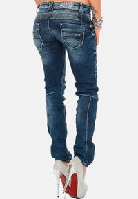 Un jean slim en denim bleu foncé présente des coutures blanches contrastées et deux poches arrière. Accompagné de talons aiguilles argentés.